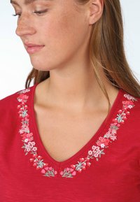 Rotes, kurzärmliges Oberteil mit V-Ausschnitt, das mit floralen Stickereien in Pink und Grün am Kragen verziert ist. Weicher Stoff und lässiges Design.