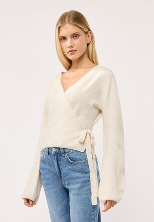 Giovane donna che indossa un maglione avvolgente color crema legato di lato e jeans blu, in piedi davanti a uno sfondo chiaro e uniforme.