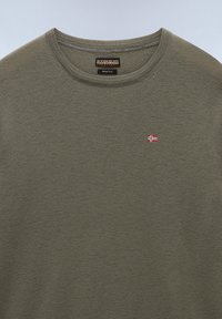 Olivgrüner Merinowollpullover mit Rundhalsausschnitt, ausgestattet mit einem kleinen roten und weißen Flaggenemblem auf der Brust sowie einem strukturierten Strickmuster.