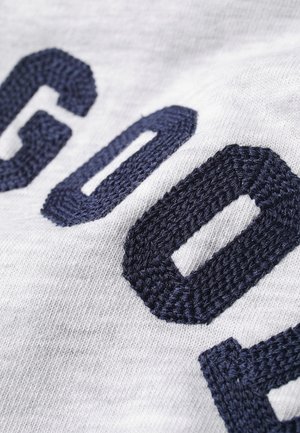 Zbliżenie na granatowo haftowane litery na jasnoszarym materiale, ukazujące teksturę i detale szwów.