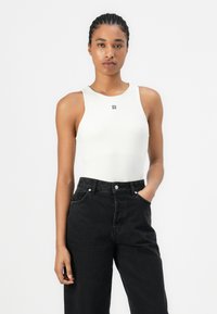 Hvit ermeløs body med rund hals og en liten svart logo, kombinert med høyhalsede svarte denimjeans. Glatt tekstur.