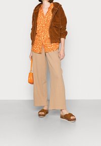 Bruine corduroy zip-up jas, oranje patroon shirt, beige wijde broek, bruine platform sandalen met knoop detail en een oranje handtas.