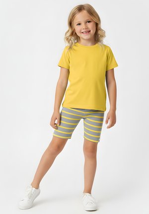 Uśmiechnięta młoda dziewczyna o blond włosach, ubrana w żółtą koszulkę, szaro-żółte paski na szortach i białe tenisówki, stojąca na jednej nodze.