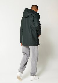 Dark green waterproof jacket with a hood, featuring a straight cut and long sleeves. Worn over light gray loose-fit pants and white sneakers.