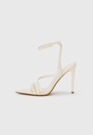 Beige sandaler med høje hæle og spids tå, med tre stropper foran og en tynd ankelrem med spænde. Glat tekstur, minimalistisk design.