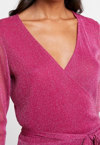 Vestido envolvente fucsia con escote en V profundo, mangas largas de tejido transparente, tela brillante y texturizada, y detalle de lazo en la cintura.