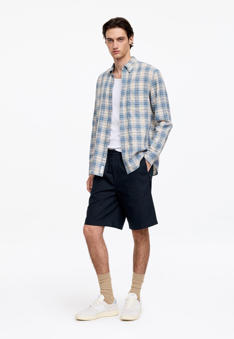 Jeune homme debout portant une chemise à carreaux bleue et beige par-dessus un débardeur blanc, un short noir, des chaussettes beiges et des baskets blanches.