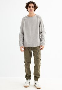 Suéter gris de punto con cuello redondo, combinado con pantalones cargo verdes oliva que tienen bolsillos con cremallera y puños elásticos, llevado con zapatillas blancas.