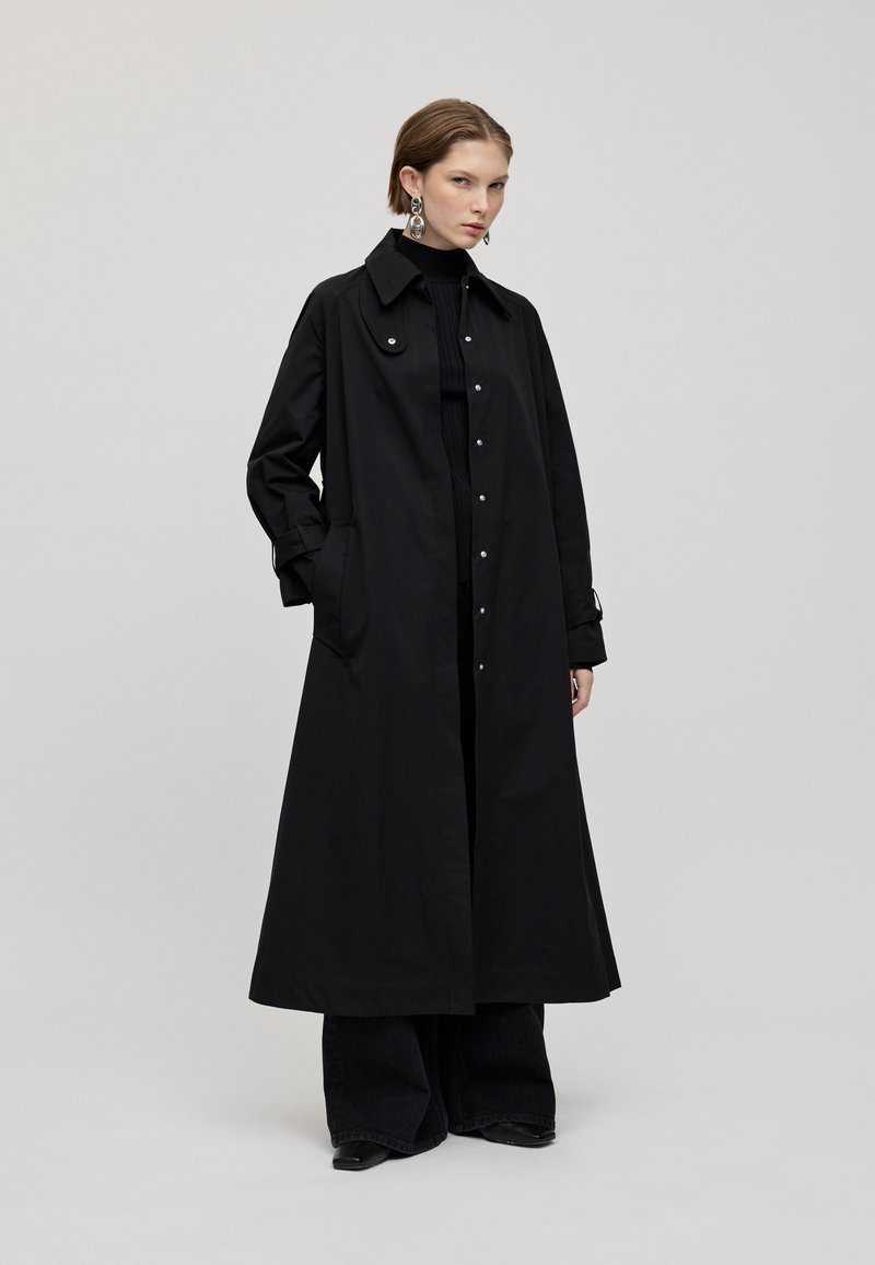 Schwarzer langer Mantel mit hohem Kragen, Druckknöpfen und seitlichen Taschen. Hergestellt aus glattem Material, mit lässiger Passform und minimalistischem Design.