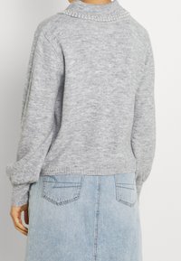 Femme portant un pull en maille gris clair avec un bord en perles sur le col et une jupe en denim bleu clair, vue de dos.
