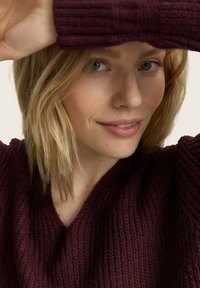 Pull en tricot bordeaux avec un col en V et une texture côtelée, porté par une personne aux cheveux blonds mi-longs, partiellement obscurcie par une main.