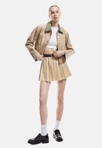 Khaki-gekürzte Jacke mit grünem Kragen, kombiniert mit einem beigen gefältelten Minirock und schwarzen Schuhen. Modell trägt weiße Socken.