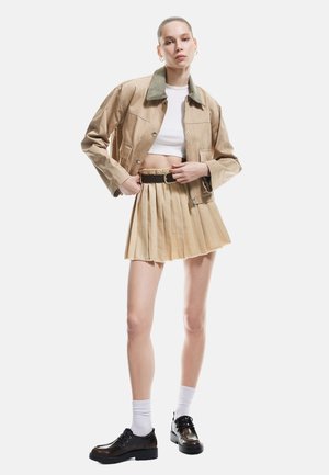 Khaki-gekürzte Jacke mit grünem Kragen, kombiniert mit einem beigen gefältelten Minirock und schwarzen Schuhen. Modell trägt weiße Socken.