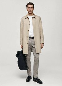 Beige knäkappor, vit skjorta, grå byxor, svarta skor. Modellen håller en svart ryggsäck och står mot en enfärgad ljus bakgrund.