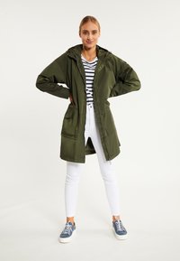 Parka impermeabil de culoare verde măslin, cu fermoar în față, glugă ajustabilă și buzunare laterale, purtat peste o cămașă cu dungi și pantaloni albi.