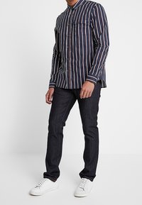 Marinblå randig skjorta med lång ärm och knäppkrage, med vita och rostfärgade ränder. I kombination med mörka jeans och vita sneakers.