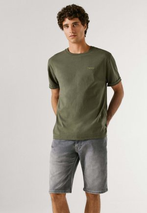 Noor mees seisab käed selja taga, kandes oliivrohelise värvi t-särki ja halli teksapükstega lühikesi pükse neutraalsel taustal.