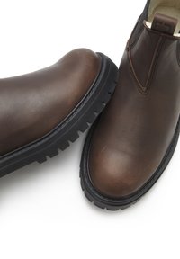 Braune Leder-Stiefeletten mit runder Zehenpartie, schwarzer Gummisohle und seitlichen elastischen Einsätzen. Glatte Textur mit minimalen Nähdetails.