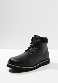 Bota de tobillo de cuero negro con un acabado suave, punta redonda, parte delantera con cordones y suela de goma con un ligero detalle en el borde. Acentos de ante en el cuello.