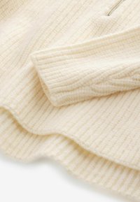 Pull en maille ivoire avec une texture côtelée, un col rond lisse et un détail de fermeture éclair au niveau du col. Matériau doux et chaud.