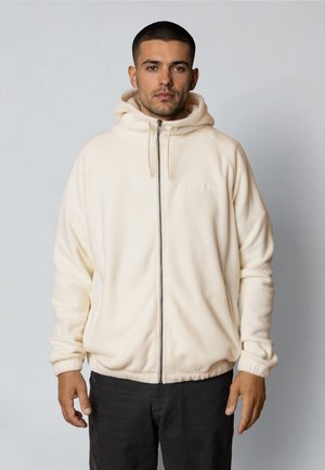 Giovane uomo con capelli corti e scuri che indossa una giacca con cappuccio color crema con cerniera e pantaloni neri, in piedi contro uno sfondo semplice.