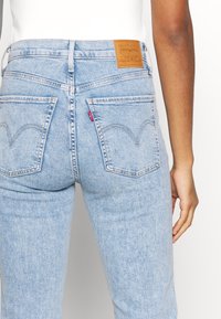 Vaaleansiniset farkut, joissa on korkeavyötäröinen malli, kaksi takataskua, ruskea nahkalappu vyötäröllä sekä hienovaraiset ompeluohjeet.