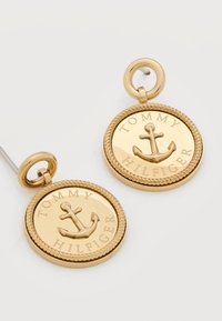 Στρογγυλά σκουλαρίκια καρφωτά από χρυσό με σχέδιο άγκυρας και την επιγραφή "Tommy Hilfiger" γύρω από την άκρη, σε λευκό φόντο.