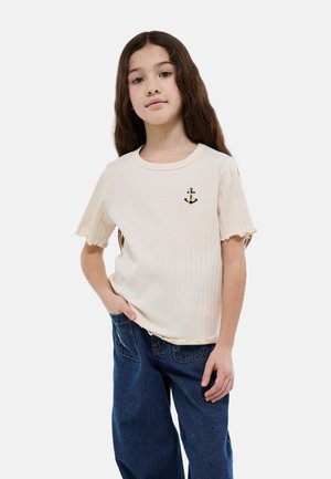 Fille aux longs cheveux bruns portant un t-shirt crème à manches courtes côtelé avec un emblème d'ancre et un jean bleu ample, debout avec une main dans la poche.