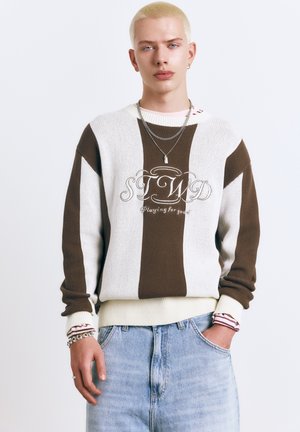 Junge Person mit kurzen blonden Haaren, die einen braun-weiß gestreiften Pullover mit dem Aufdruck "STWD Playing for good" und hellblaue Jeans trägt.