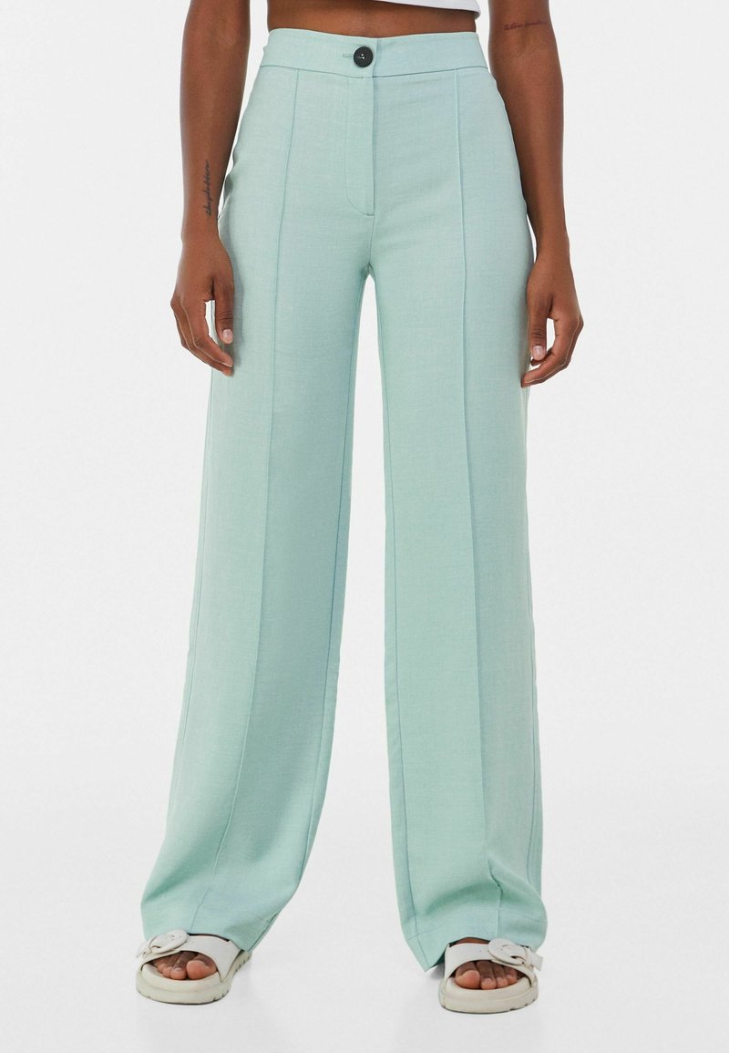 Personne portant un pantalon large taille haute bleu clair avec un bouton à l'avant et des sandales blanches ouvertes sur fond blanc.