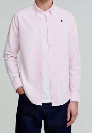Homme portant une chemise rose clair boutonnée sur un t-shirt blanc et un jean foncé, la main gauche dans la poche et un petit logo de crâne sur la poitrine.