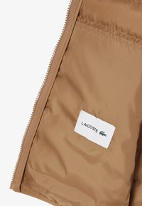 Beige pufferjack binnenkant met een gladde afwerking; voorzien van een zichtbare rits en een label met "LACOSTE" en logo.