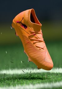 Leuchtend orangefarbener Fußballschuh mit strukturierter Oberseite, auffälligen Schnürsenkeln und einem schwarzen Logo. Auf Gras mit verstreuten Halmen erhoben.