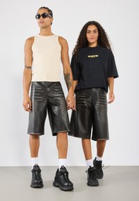 Dva modelky v ležérnych outfitoch. Jedna má na sebe béžový rebrovaný tielko a čierne šortky z umelého kože, druhá má čiernu košeľu a zodpovedajúce šortky.