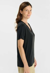 Vrouw met lang bruin haar, gekleed in een zwart V-hals kortemouwen top en beige broek, staande en zijwaarts kijkend tegen een witte achtergrond.