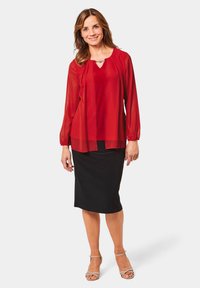 Rote transparente Bluse über einem roten Top, kombiniert mit einem schwarzen Bleistiftrock. Hat lange Ärmel und ein dezentes Dekolleté. Beigefarbene Sandalen.