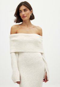 Abito maglione off-the-shoulder realizzato in morbido materiale a maglia color crema. Presenta dettagli a coste e maniche lunghe con aperture ai polsi.