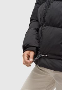 Zwarte gewatteerde jas met een ritssluiting aan de voorkant, elastische manchetten en een verstelbare trekkoordzoom. Glad materiaal met gewatteerde delen voor isolatie.