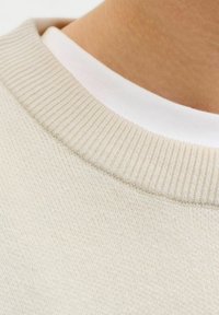 Gros plan d'une personne portant un pull en maille beige sur un t-shirt blanc à col rond, montrant le col et la peau.