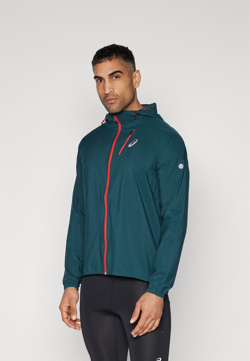Giacca leggera color teal con cappuccio, caratterizzata da zip e dettagli rossi, tasca sul petto con zip e polsini elasticizzati. Realizzata in un materiale liscio e resistente all'acqua.