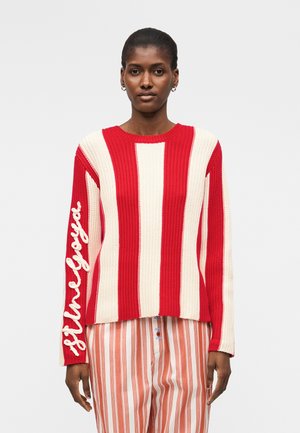 Žena nosi červeno-biely vertikálne pruhovaný pletený sveter s nápisom „Still Loyal“ na rukáve a červeno-biele pruhované nohavice.