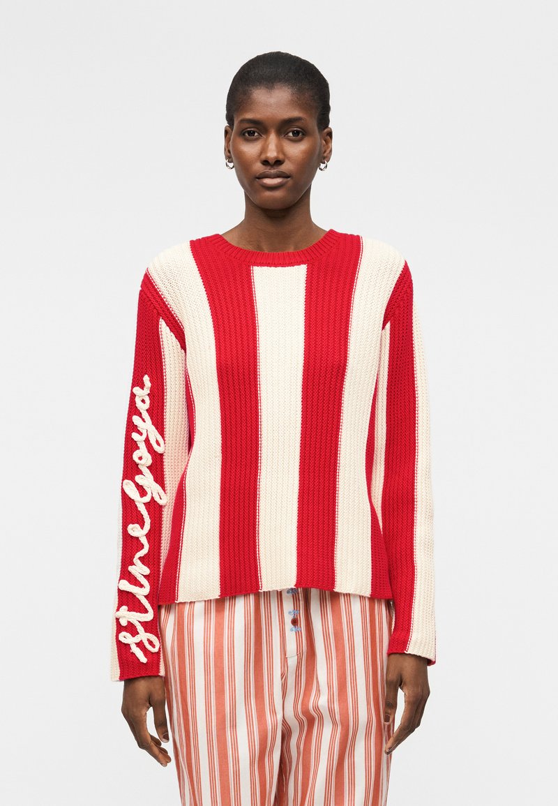 Γυναίκα φοράει κόκκινο και λευκό πλεκτό πουλόβερ με κάθετες ρίγες και τη φράση "Still Loyal" στο μανίκι και παντελόνι με κόκκινες και λευκές ρίγες.