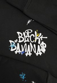 Zwarte stof met witte graffiti-stijl tekst "Black Bananas" en kleine geborduurde verfspetters in blauw, geel en wit rondom de letters.