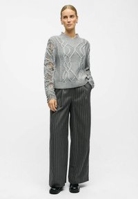 Pull en tricot gris avec des motifs en relief complexes et des manches en dentelle transparente. Associé à un pantalon à jambes larges à rayures et des chaussures noires.