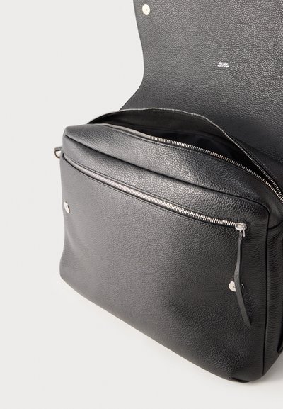 Μαύρη τσάντα από δέρμα με υφή, με εμπρόσθια τσέπη με φερμουάρ και ανοιχτό επάνω καπάκι που αποκαλύπτει το άδειο εσωτερικό, σε απλό φόντο.