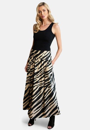 Žena s dlhými blond vlasmi, oblečená v čiernom tielku bez rukávov, v maxi sukni s vysokým pásom so zebrovým vzorom a v čiernych členkových topánkach s otvorenou špičkou, stojaca.