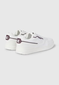Białe sneakersy z teksturowanym fioletowym akcentem na pięcie, wyściełanym językiem oraz gumową podeszwą z poziomymi rowkami dla lepszej przyczepności.