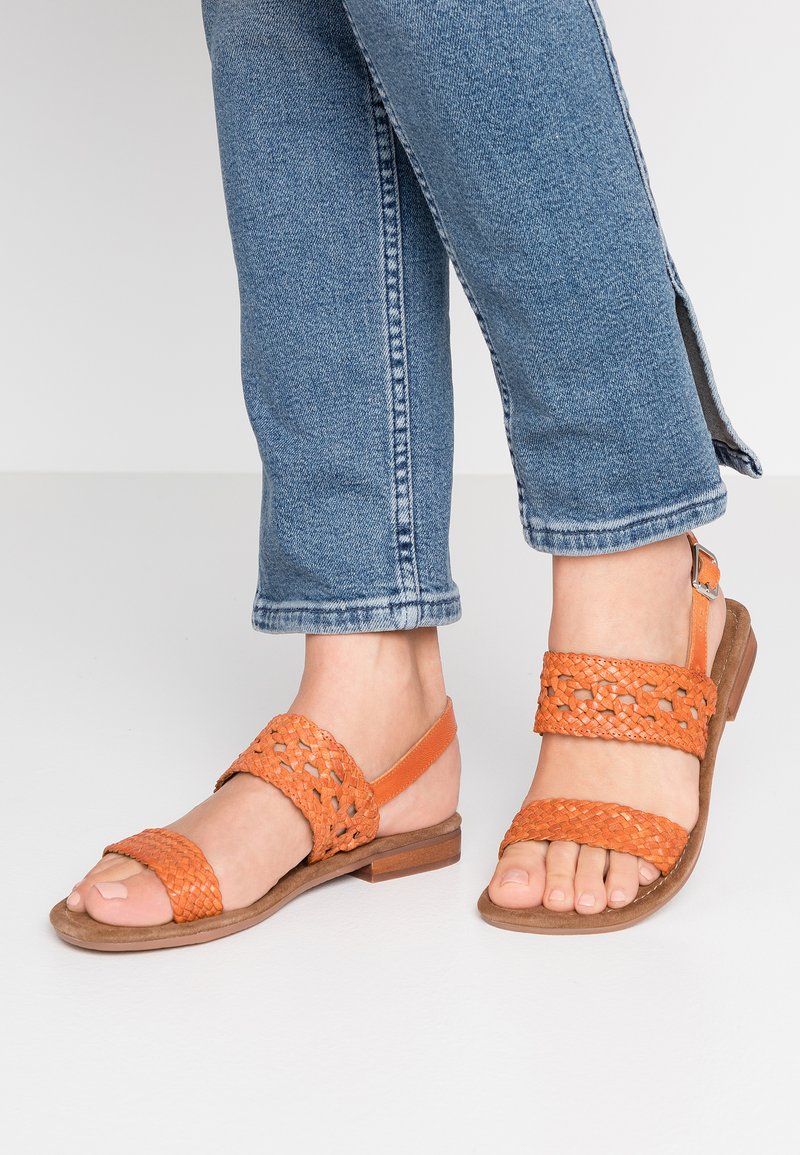 Orange woven sandals with open toe and two wide straps, set on a brown flat sole. Worn with slightly cropped blue denim pants.