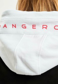 Bijela dukserica s crvenim natpisom "DANGER" na leđima. Mekani pamučni materijal, s oblikovanom kapuljačom i kontrastnim crnim tijelom.