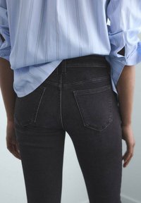 Persona vistiendo jeans de mezclilla negros y una camisa de rayas azul claro con las mangas arremangadas, vista desde atrás a la altura de la cintura.